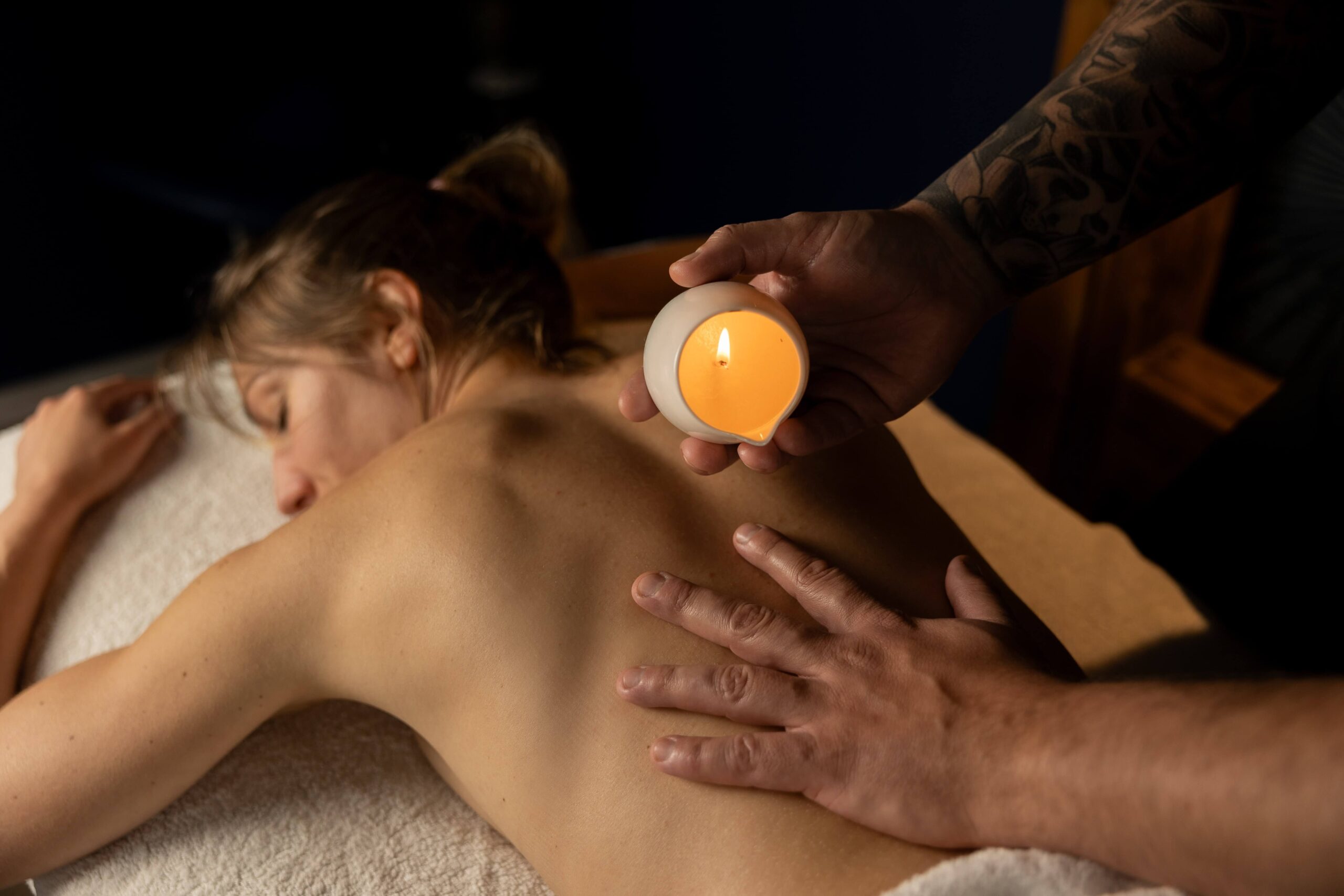 Zdjęcie ukazuje parę leżącą obok siebie na stołach do masażu w gabinecie Oriental SPA w Jeleniej Górze, podczas masażu ciepłą świecą. Masażyści delikatnie wlewają ciepły, aromatyczny wosk ze świec na plecy pacjentów, wykonując relaksacyjne ruchy. Atmosfera gabinetu jest romantyczna i spokojna, sprzyjająca głębokiemu odprężeniu i wspólnemu relaksowi pary.
