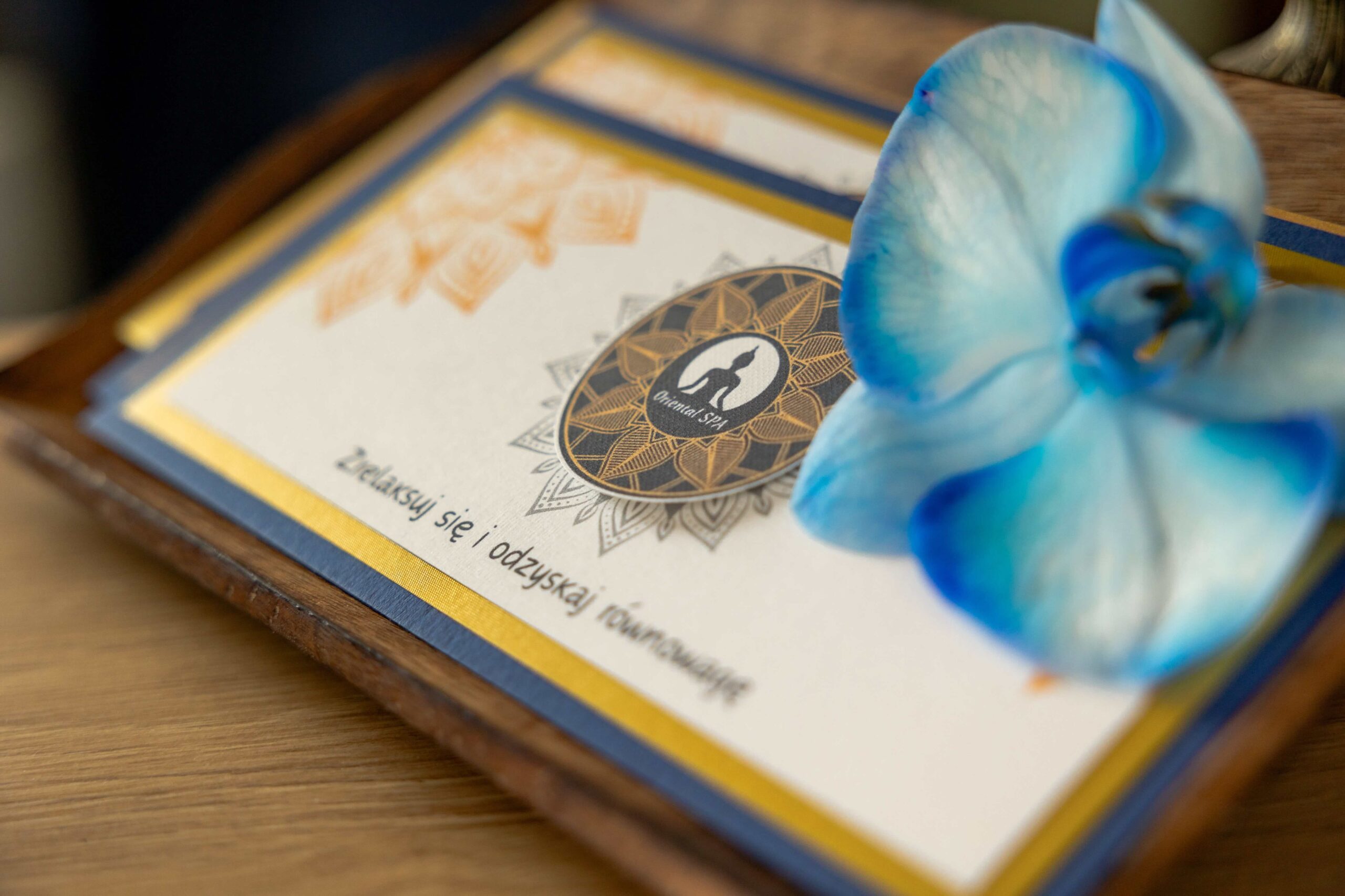 Zdjęcie przedstawia elegancki bon podarunkowy na masaż. Voucher wykonany jest na wysokiej jakości papierze, zdobiony delikatnymi wzorami oraz estetycznym logo gabinetu masażu. Na pierwszym planie widoczne są czytelnie napisane informacje, takie jak "Bon podarunkowy" oraz "Voucher na masaż", a także dane kontaktowe i adres gabinetu. W tle można dostrzec relaksacyjne elementy, takie jak świece, kamienie do masażu, kwiaty i ręczniki, które tworzą przyjemną i odprężającą atmosferę. Całość utrzymana jest w ciepłych, uspokajających kolorach, podkreślających luksus i wyjątkowość podarunku.